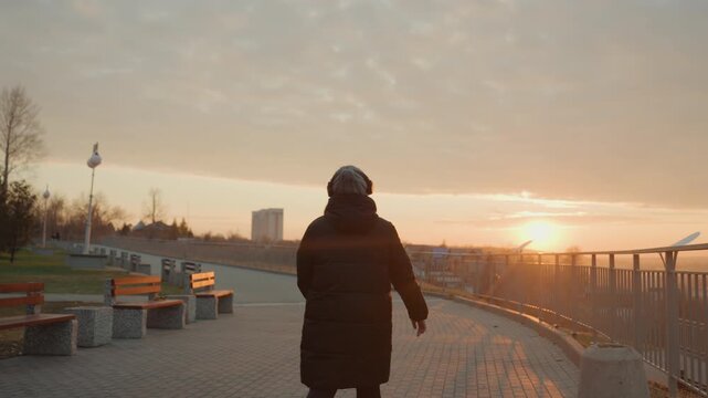 Silhouette walking toward sunrise dancing, arms outstretched on empty promenade, golden horizon glow, benches and railing along riverfront, headphones on, rhythmic steps and joyful motion, morning