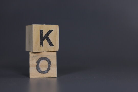 Two wooden blocks stacked vertically with the letters "K" and "O" printed in black, forming the word "KO" against a dark grey background, symbolizing typography, and simple creative concept.