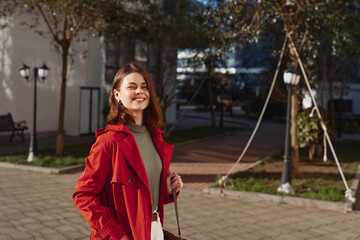 Obraz premium Young woman in a red coat outdoors, smiling under afternoon light, walking in a sunny plaza with trees and modern buildings, a soft breeze rustling her hair for a candid lifestyle moment
