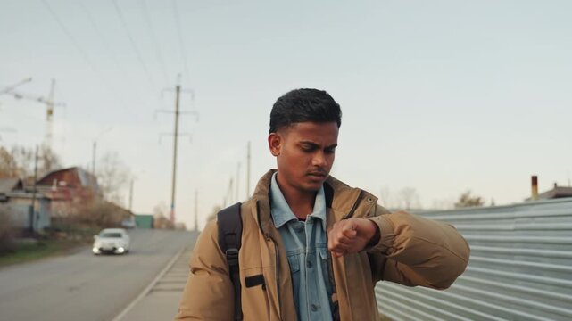 Man on roadside repeatedly checking watch and glancing at passing car, metal fence and cranes line street, jacket and backpack visible, tense hurry and urgency suggesting imminent repair task