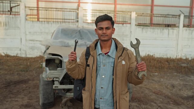 Indian man demonstrating wrenches outdoors, standing before wrecked vehicle at salvage yard holding adjustable wrench and pliers, confident instructor energy with denim shirt and layered jacket,