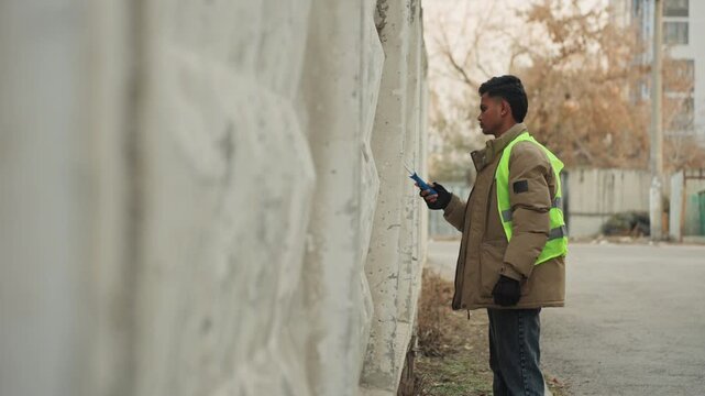 Worker in reflective vest inspects wall for repair planning, documenting cracks with tablet, assessing concrete spalling and structural condition, cold urban alley, gloves and jacket, methodical
