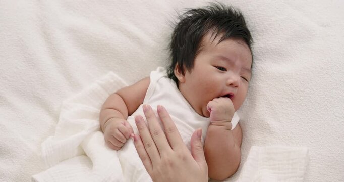 Asian infant boy lying on white blanket while yawning and sucking fist, with gentle mother touch expressing sleepiness, warmth, and nurturing care in peaceful early childhood moment