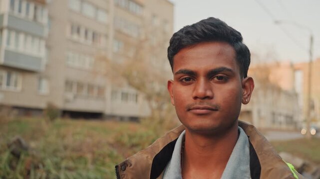Young south asian man in jacket near apartment building, contemplative repair apprentice inspecting facade, maintenance worker checking surroundings, closeup portrait in autumn light, reflective mood