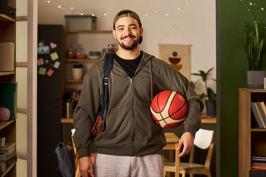 Portrait of young adult biracial man carrying basketball and bag, smiling at camera in home interior, representing student lifestyle and active routine. Useful for education or youth marketing