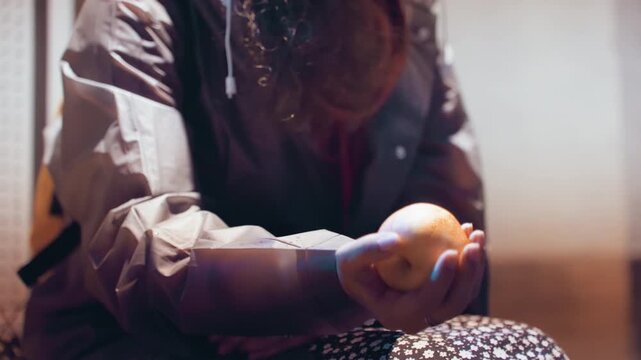 Soft indoor hand balancing orange closeup, seated woman adjusting fruit with fingertips, patterned skirt and jacket, warm window glow, candid motion study, gentle toss and catch, contemplative