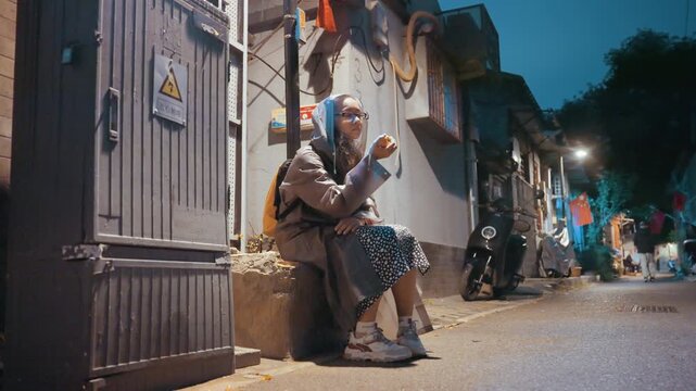 Elderly woman sitting on doorway under warm streetlight in narrow urban alley, backpack at her side, motorbikes passing, quiet dusk mood, contemplative expression, textured walls and metal gate,