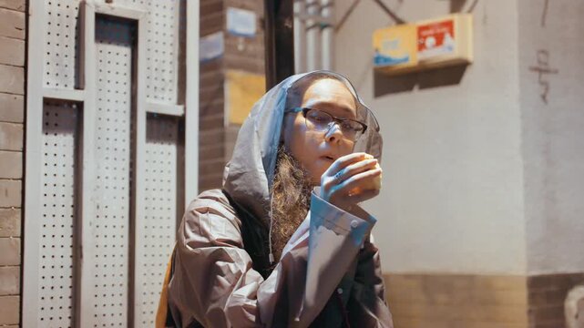 Urban street portrait curious observer adjusting glasses and scarf, hooded figure leaning on metal grate, chewing snack and scanning neonlit lane, candid tactile texture, quiet tension and gentle hum