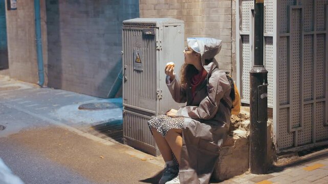 Asian woman sitting on sidewalk under lamp, tired commuter resting against utility box in hooded coat, hands clasped, vintage film tones, melancholic nighttime glow and wet asphalt texture