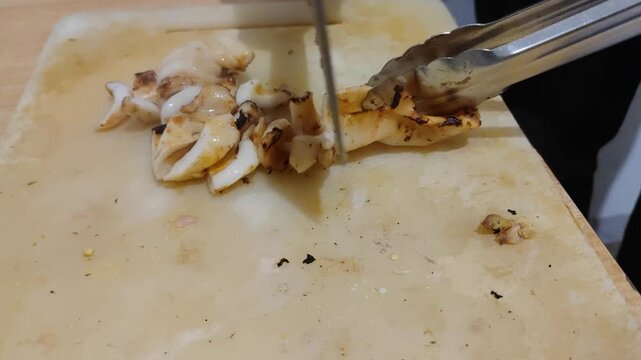 Close-up video of freshly grilled squid being sliced on a cutting board using kitchen tongs and scissors. Delicious seafood preparation process, perfect for culinary, street food, and cooking concept 