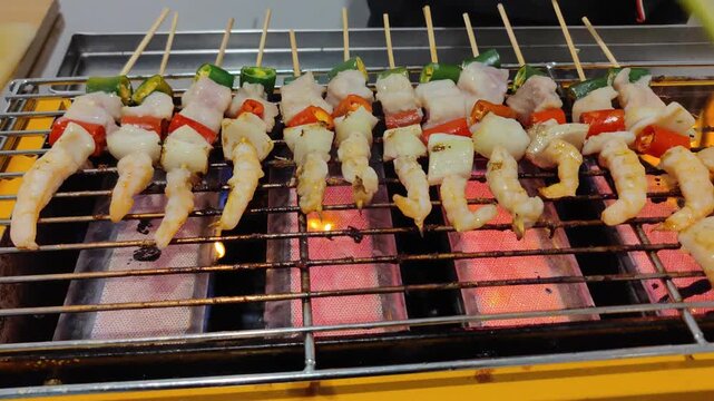 Close-up video of shrimp skewers being grilled over charcoal fire with vegetables such as chili and green onions. Delicious seafood street food, smoky barbecue atmosphere, perfect for culinary