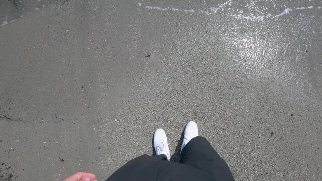 Point of view shot from above of a person in white sneakers standing on wet sand, sunlight reflecting on beach as calm waves roll in. Real-time, natural daylight, handheld camera motion.