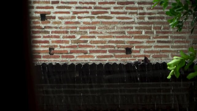 Handheld close up of heavy rain water flowing and dripping from a corrugated zinc roof of a village house with a brick wall background during monsoon season in East Java, Indonesia.