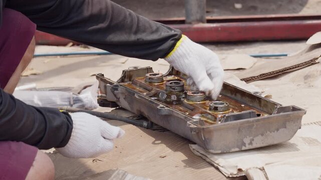 Hands inspect worn engine block. White gloves touch metal surfaces carefully. Workshop floor holds tools and debris. Person focuses intently on mechanical repair