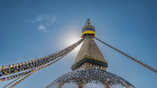 A huge stupa. The eyes of the Buddha are painted on the top. A spire against a clear blue sky. Rows of bright multicolored flags. A flock of pigeons on the dome. Nepal. Kathmandu. Boudhanath Stupa