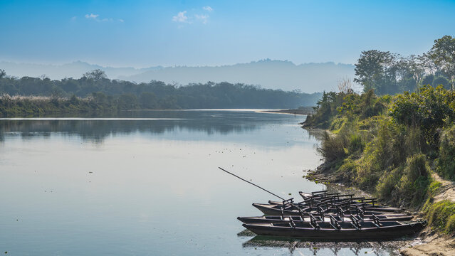 Wooden boats are moored at the riverbank in the jungle. Green trees on the shore. Reflection on calm water. Mountains in the distance against the blue sky. Nepal. Chitwan National Park. Rapti river.