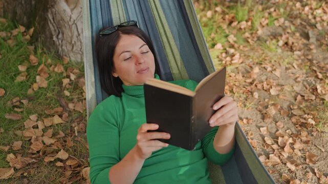 White woman hammock camping reads aloud, expressive gestures and dramatic narration, green sweater, fallen leaves underfoot, sunglasses perched, voice carrying through forest, performer energy,