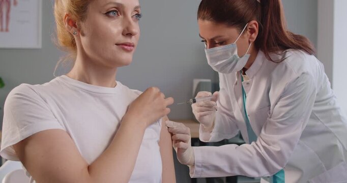 Immunization of adult woman in hospital for flu and coronavirus prevention, doctor in gloves giving shot of vaccine to patients arm. Girl sitting, waiting for vaccination with trust, nurse injecting