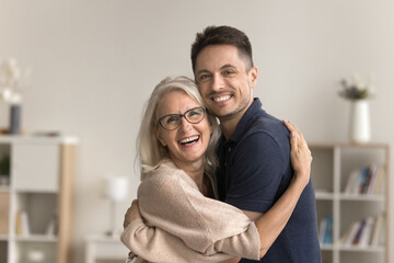 Fototapeta premium Beautiful mature woman cuddling her young adult son, laughing, pose in domestic room, look for camera with positive, joyful expression. Family attachment, love, support. Happy Mothers Day celebration