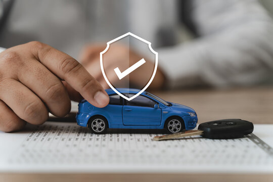 Auto insurance coverage and vehicle protection concept with agent hand presenting blue car model on policy contract document beside car key and shield checkmark icon on office desk surface.