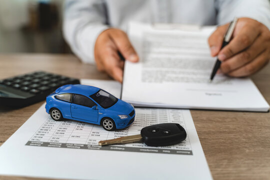 Hands reviewing car loan finance document contract with payment schedule table, blue toy car model and key on paperwork, calculator on desk, representing auto finance agreement analysis, vehicle lease