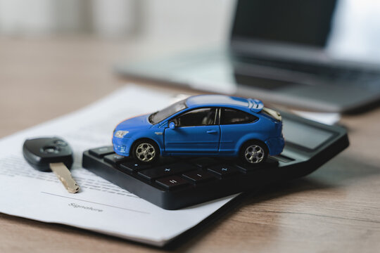 Miniature blue vehicle model positioned on finance calculator with key and purchase contract showing signature area, laptop workspace background, representing automotive pricing estimation, loan cost 