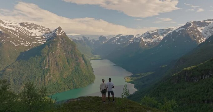 Couple is standing at breathtaking viewpoint looking out over the turquoise fjord waters of Oldevatnet, snowcapped peaks and Briksdalsbreen glacier in the distance under a blue sky, Klovane hike