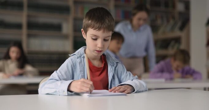Focused Caucasian schoolboy child making schoolwork, writing in copybook during lesson sitting at desk in classroom. Concept of primary school education, learning process, early skill development