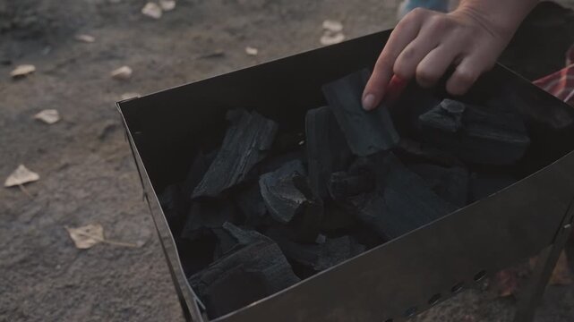 Cold charcoal in portable grill camping, closeup of stacked black coals inside metal brazier on dirt ground with scattered leaves, dusk light casts muted tones, smoky atmosphere signals campfire