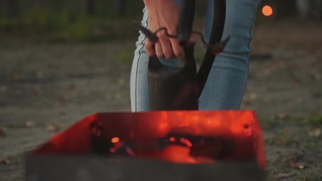 Camper pouring liquid into glowing embers, jeansclad legs and hands holding metal kettle with strap, sparks lifting from rectangular firepit, evening campsite maintenance and safety action captured