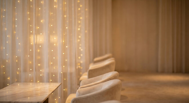 A cozy seating area with plush chairs and a marble table is illuminated by soft string lights draped behind sheer curtains, creating a warm and inviting atmosphere.