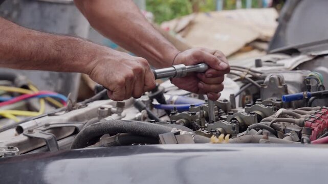Hands tighten engine bolts with a torque wrench. Fingers grip tools firmly, showing focused effort. Mechanic works under open hood, surrounded by wires and metal. Scene captures hands-on repair