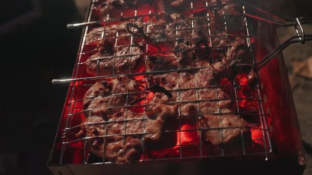 Closeup camping grill sizzling steak over glowing coals, marinated meat on wire rack, tongs searing edges, warm ember glow and ash, intimate nighttime camp cook preparing rustic dinner