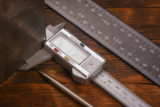 Digital caliper measuring metal sheet with steel ruler and metal scribe on wooden workbench table close up background.