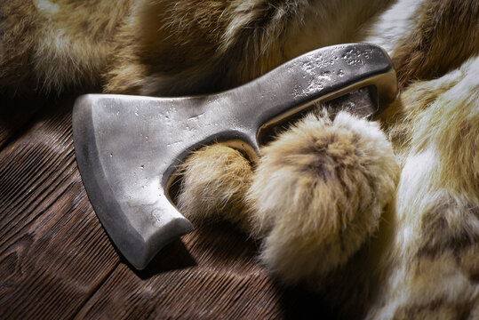 Forged medieval axe head and animal fur placed on dark wooden workbench table top view background.
