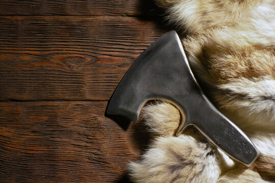 Forged medieval axe head and animal fur placed on dark wooden workbench table top view background.