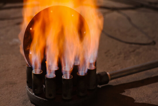 Gas metal heating burner with multiple nozzles producing blue and orange flames heating round steel plate on workshop floor background.