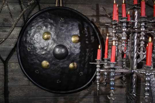 Forged black steel viking round shield with central boss mounted on dark wooden wall surrounded by wrought iron candle holders with red candles and medieval weapon.