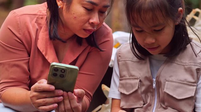 Asian mother and daughter using smartphone for digital learning and research in a park, family technology and education concept.