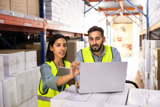 Workers working in storage aisle, using laptop and digital tablet to review inventory data, check stock levels, coordinate logistics operations. Distribution and supply chain management in warehouse
