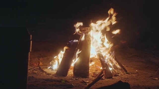 Night fire vigil. Survivalist observes intense flames and rugged surroundings carefully. Protective lookout maintains vigil over blazing logs amidst harsh shadows and survival gear in night