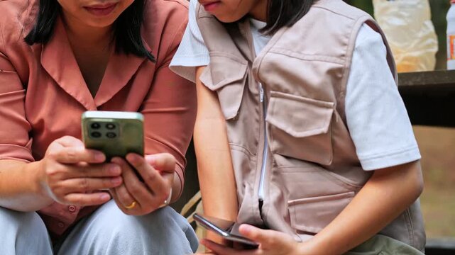 Asian mother and daughter using smartphone for digital learning and research in a park, family technology and education concept.