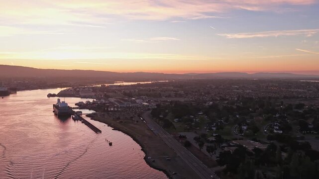 Beautiful aerial drone perspective flying over the Oakland estuary during a colorful sunrise, revealing the cityscapes of Oakland and Alameda with warm light reflecting on the water