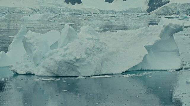 High angle parallax dolly shot glides past massive textured sculpted iceberg reflecting in calm teal Antarctic ocean with glacier behind