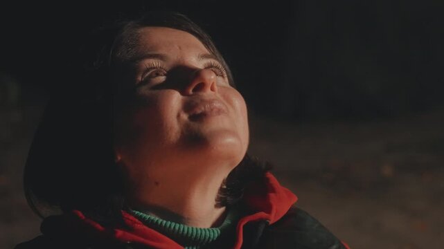 Hispanic woman by campfire at night, soft firelight illuminating face and hooded jacket, eyes closing in peaceful reflection then opening into warm smile smoky woods backdrop, intimate storyteller