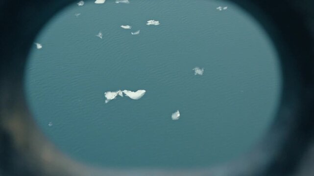 View through round expedition ship porthole looking down at teal Antarctic ocean scattered with drifting ice floes and brash ice