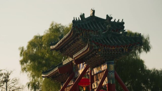 Ornate pavilion roof framed by willow, painted eaves and red pillars bathed in golden sunlight, closeup of carved timber detail, peaceful park atmosphere with quiet pathways and restoration worker