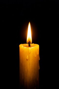 Single yellow candle burning brightly against a pitch black background. Warm glowing flame with melting wax, creating a peaceful and moody atmosphere.