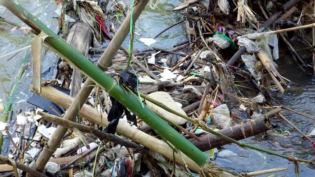 Handheld close-up of a severely polluted river with entangled plastic waste and bamboo debris at Tawang Pare dam, Kediri, Indonesia, highlighting severe environmental contamination.