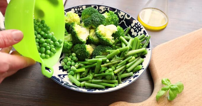 Woman hands pouring green peas from silicone colander into bowl with broccoli and asparagus. Chef prepares fresh organic vegetables for healthy vegetarian meal on wooden table. Process of mixing raw
